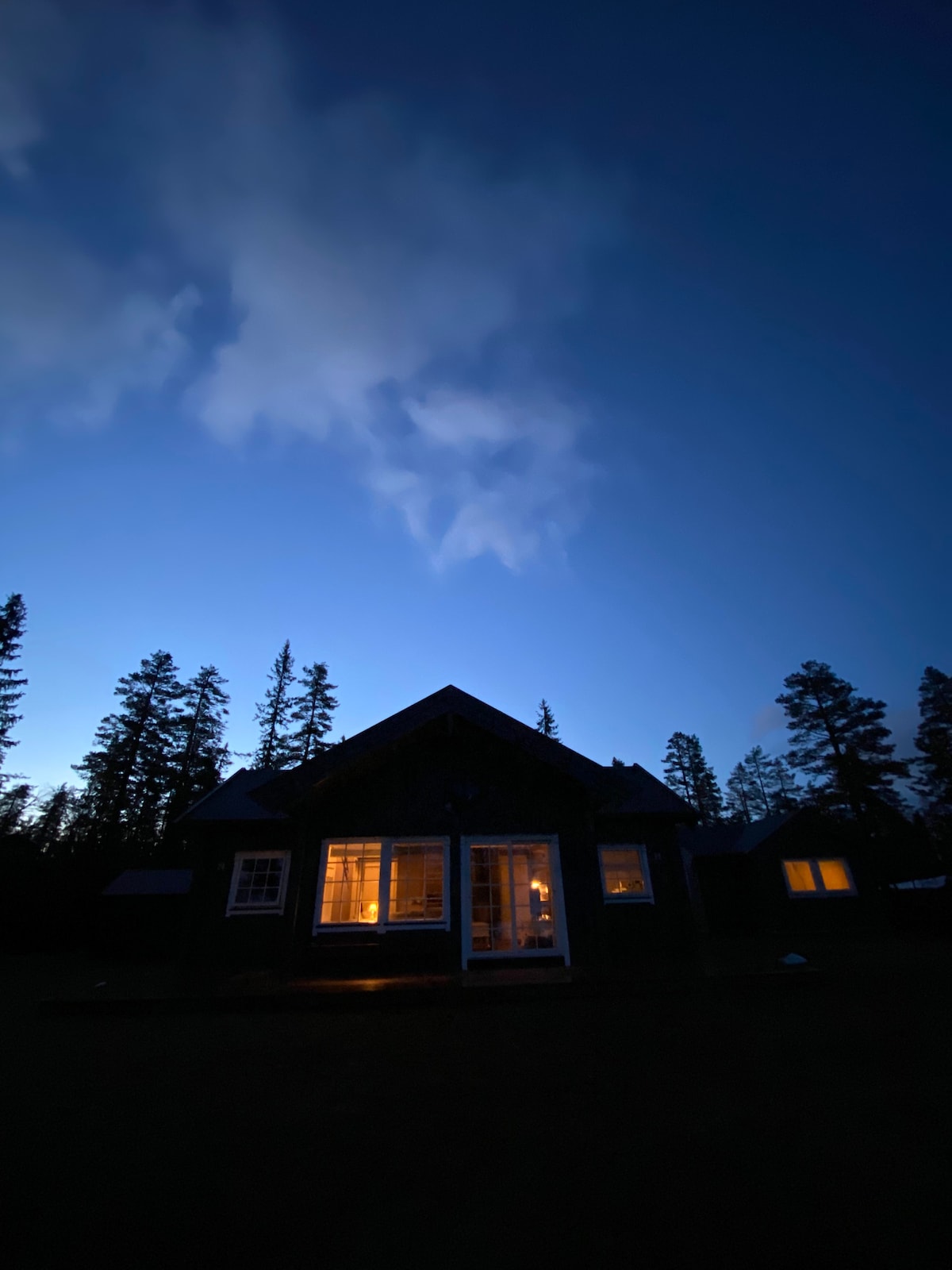 The cabin is illuminated from within, standing against a deep blue sky at dusk. Tall trees frame the scene, and wispy clouds are visible above, creating a serene wilderness atmosphere.