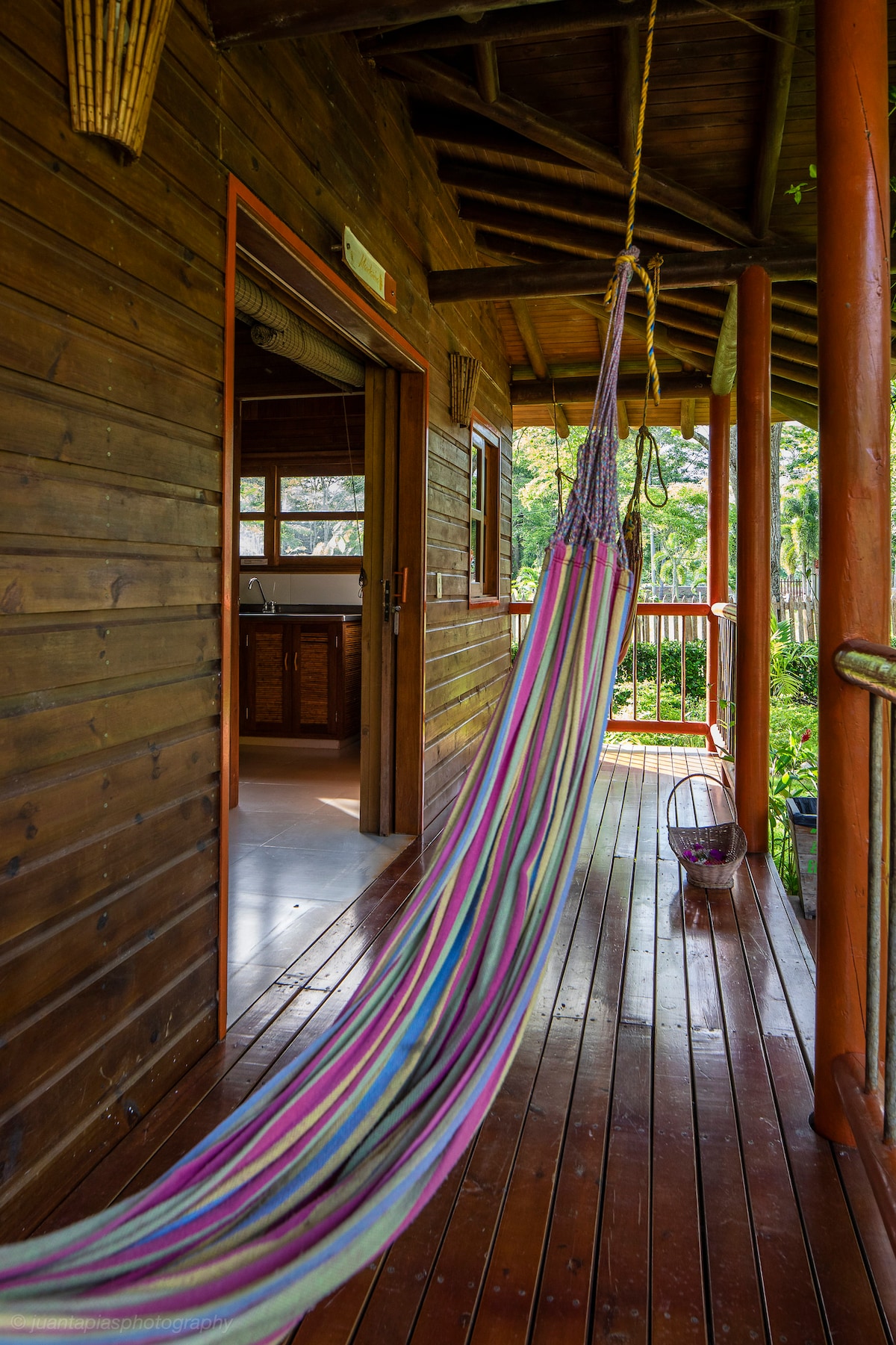 A colorful hammock is suspended from the ceiling of a wooden porch, inviting relaxation. The warm wood tones of the cabin are complemented by the vibrant stripes of the hammock, with lush greenery visible through the open doorway in the background.