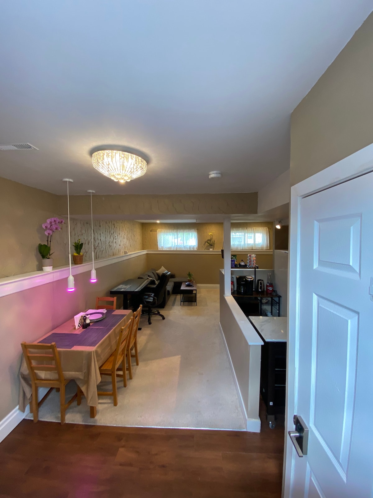 An open-plan living space features a dining area with a table set for four, complemented by overhead pendant lights. A cozy sitting area is visible with a sleek sofa and a coffee table. Soft natural light enters through large windows.