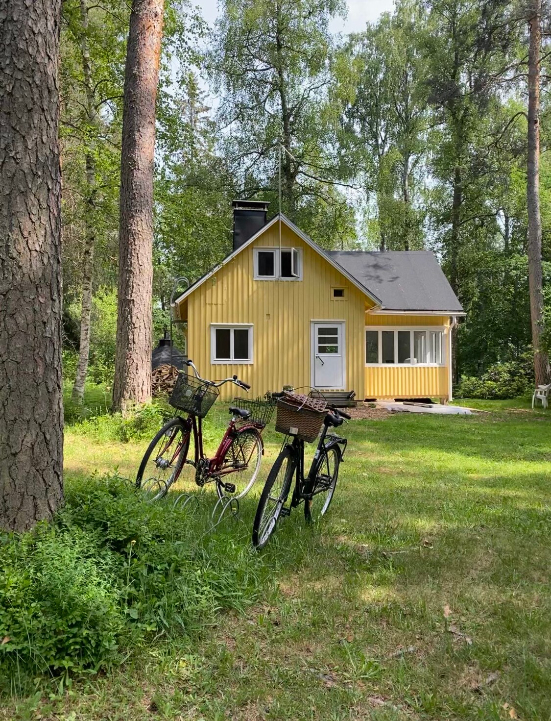 A charming yellow cottage is nestled among tall trees, creating a serene atmosphere. Two bicycles are parked on the green grass, hinting at leisurely outdoor explorations in the surrounding nature. Large windows reflect the greenery, inviting natural light indoors.