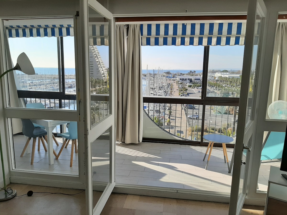 A spacious interior features large glass doors that open to a closed terrace, offering unobstructed views of the marina and sea. Natural light fills the area, complemented by striped awnings. Two light blue chairs and a small table are positioned on the terrace, creating a relaxing space.
