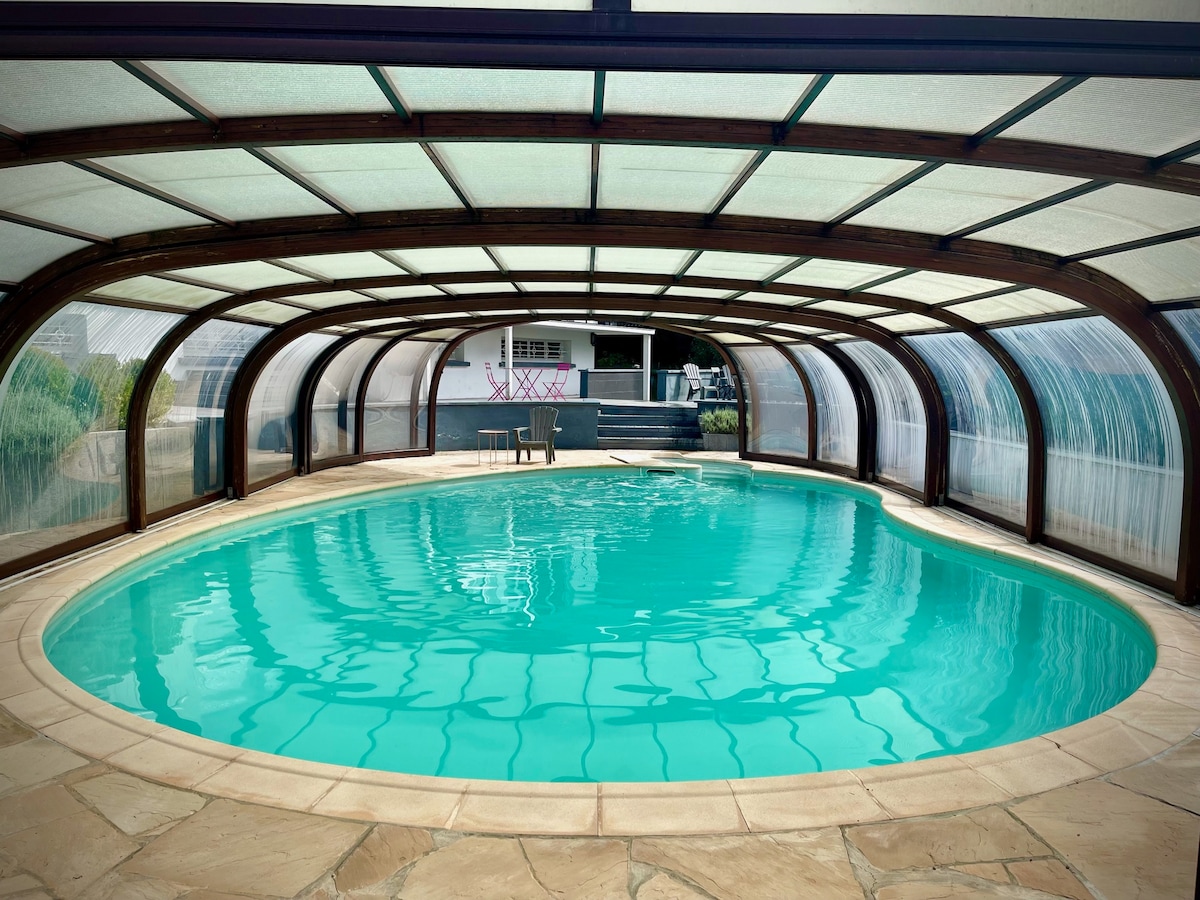 An indoor pool features a modern design with a curved glass enclosure. The water appears inviting in a bright turquoise hue, reflecting the light from the overhead structure. Surrounding the pool, a patio area is visible, complementing the tranquil setting.