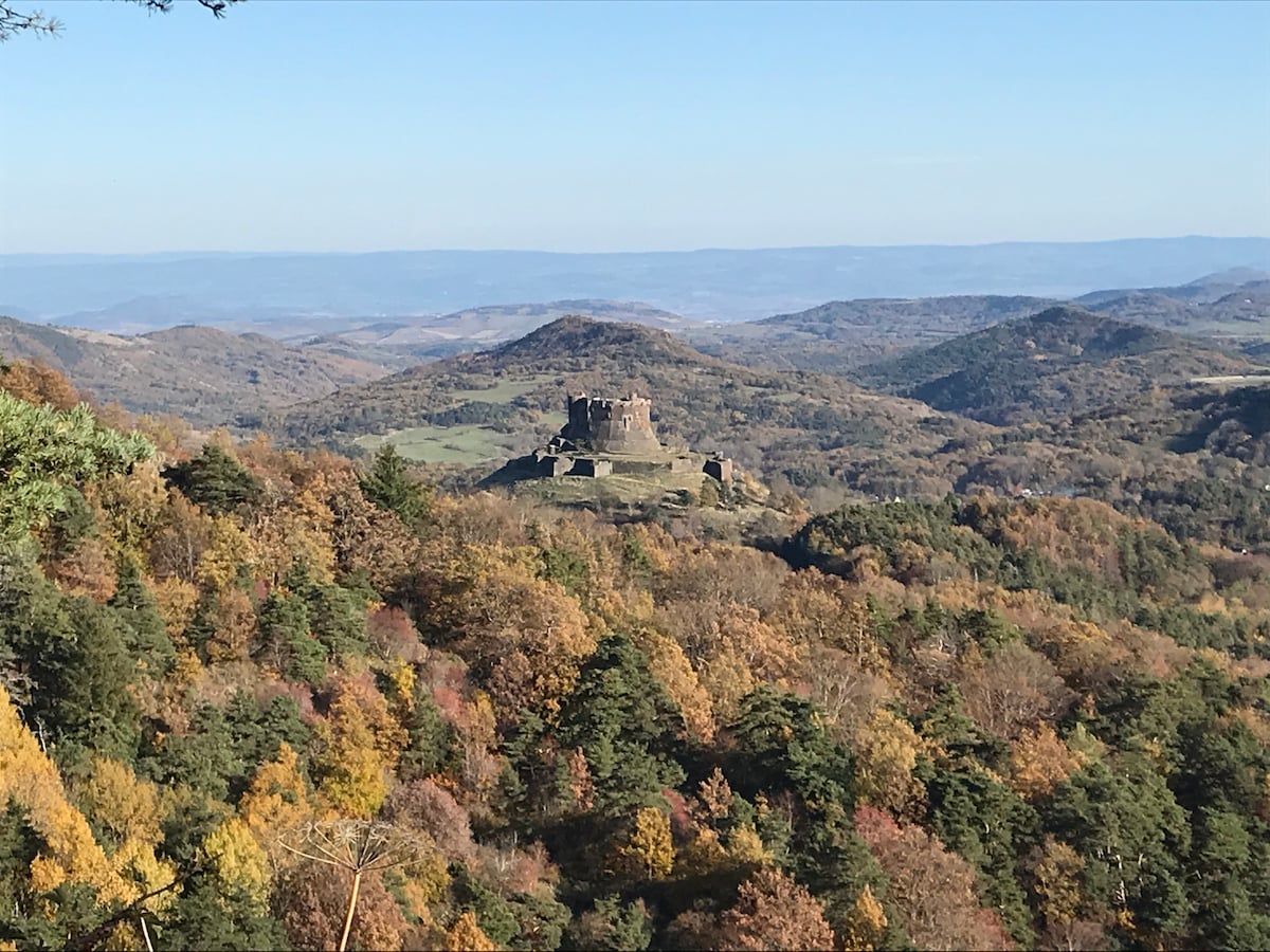 A distant view of a historic castle is framed by rolling hills and autumnal forests. The landscape displays a palette of warm fall colors, with trees showcasing vibrant oranges, yellows, and greens under a clear blue sky.