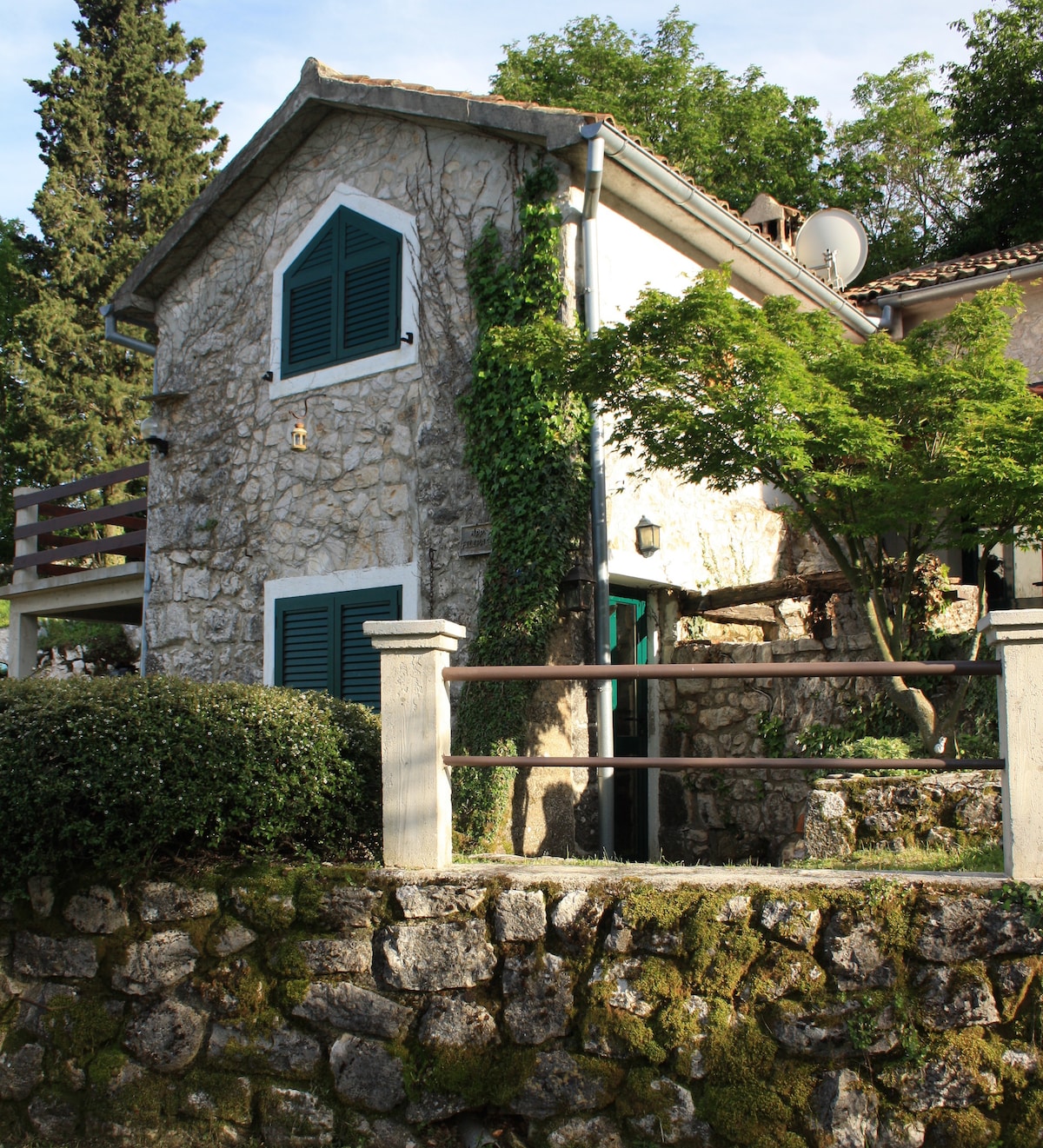 The charming stone exterior of the house is complemented by green shutters and surrounding foliage. A low stone wall encircles the garden, with a pathway leading up to the entrance. Mature trees and plants enhance the natural setting.