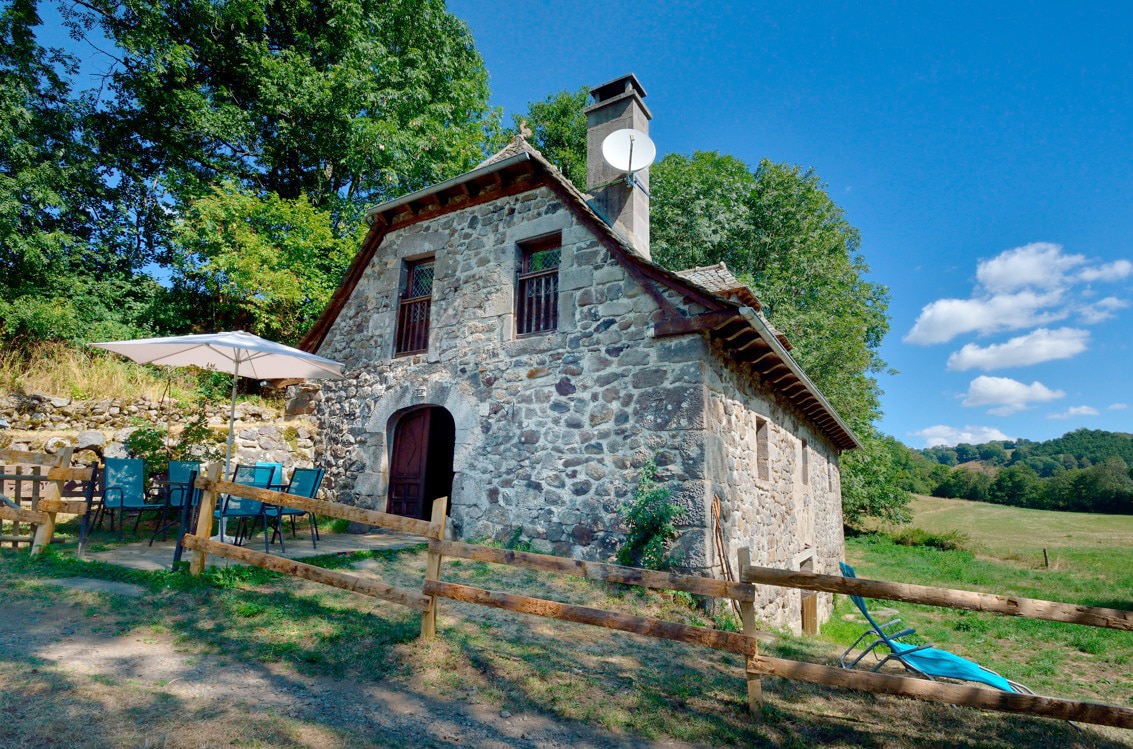 A rustic stone mill is set against a vibrant blue sky, surrounded by lush greenery. Outdoor seating is provided under a parasol, accompanied by a fenced area. A satellite dish is mounted on the roof, blending with the charming natural landscape.