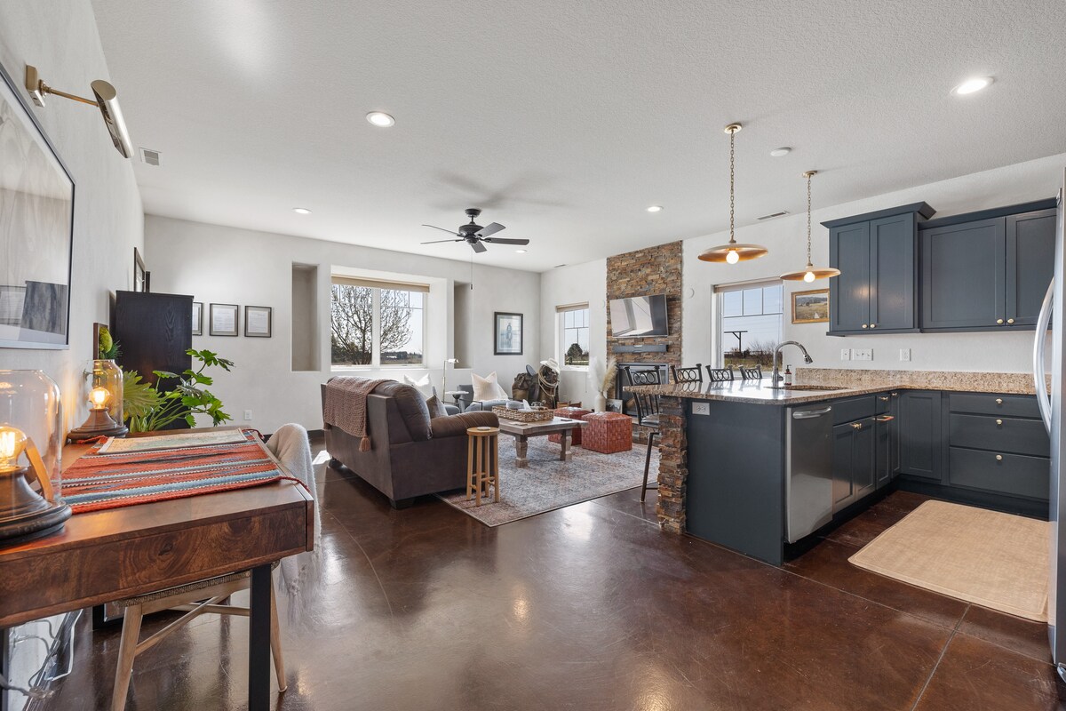 A spacious living area features a comfortable couch and a rustic stone fireplace. An inviting kitchen with dark cabinetry and granite countertops is seen, along with a dining space that includes a wooden table. Natural light filters through large windows, enhancing the open layout.