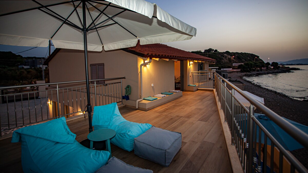 A spacious balcony features two cushioned lounge chairs and a small table, providing a relaxing space. A large umbrella offers shade, while the serene view of the beach and sea is visible in the background. Soft lighting creates a calming ambiance for evening relaxation.