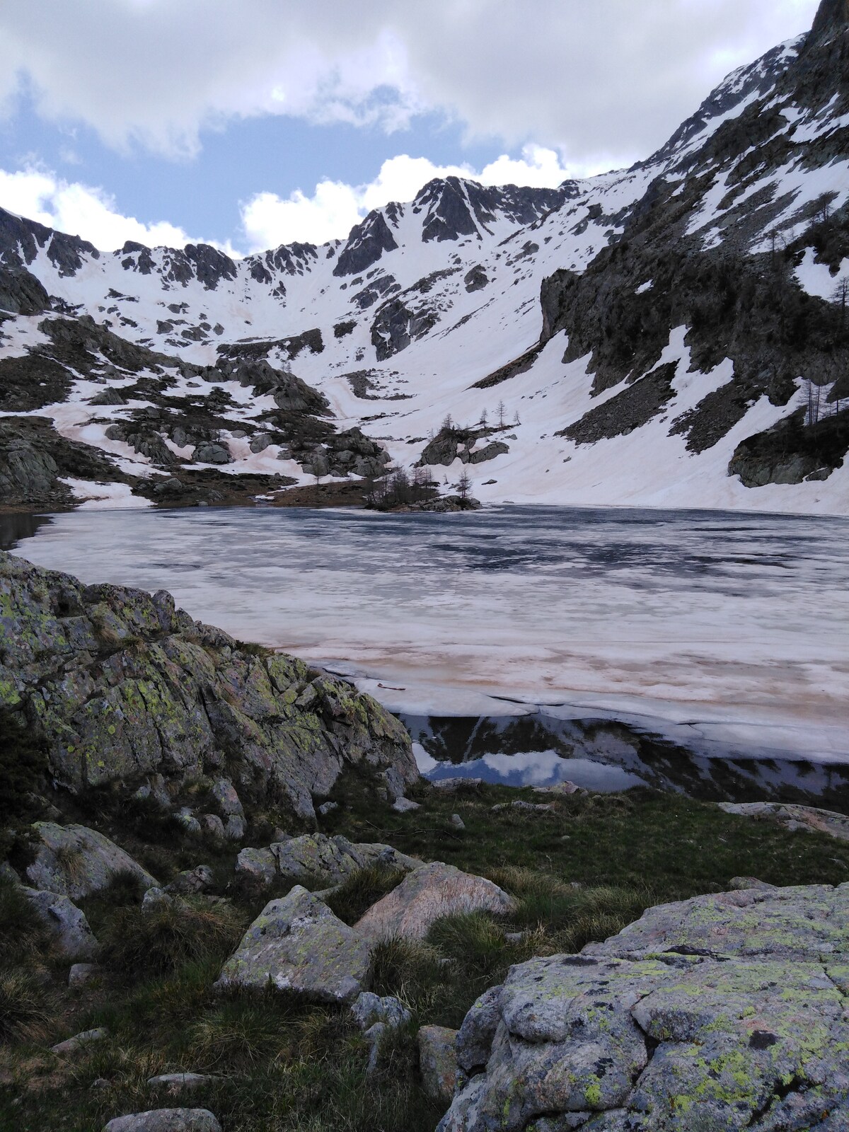 A serene landscape showcases a partially frozen lake surrounded by snow-covered mountains. Rocky terrain with patches of green grass leads to the water's edge, reflecting the rugged peaks in the background. The scene is framed by a clear sky with scattered clouds.