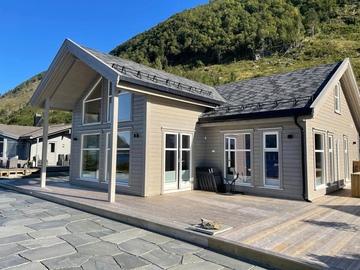 The exterior of the vacation home displays a modern design with large windows that allow plenty of natural light. The spacious deck features wooden flooring and provides access to the landscape, framed by lush green hills in the background.