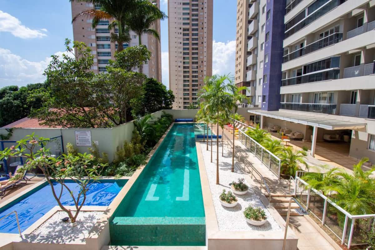 Two swimming pools are situated within a landscaped area, surrounded by palm trees and greenery. The larger pool features a sleek design with clear blue water, while the smaller pool is framed with a stone border. A shaded sitting area is visible nearby.