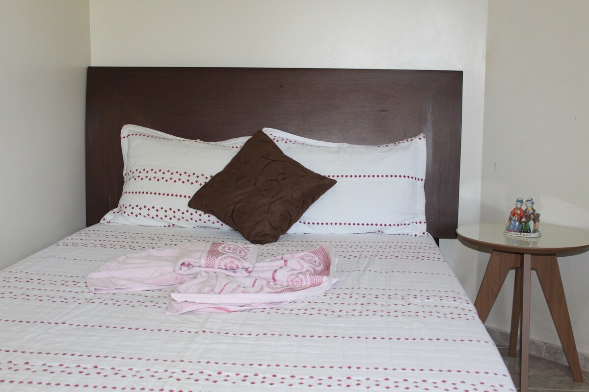 A spacious bed is presented with simple striped bedding and decorative pillows. A small bedside table holds a set of glasses. A light pink towel is casually draped over the bed, enhancing the serene ambiance of the room.