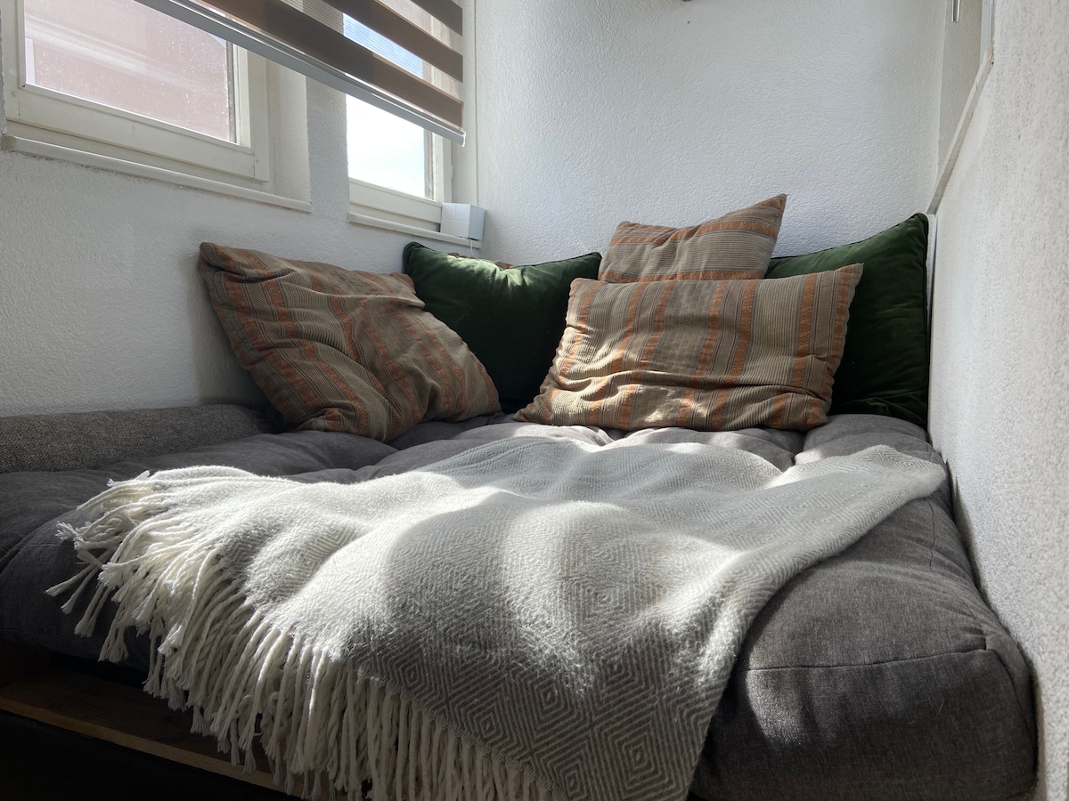 A cozy seating area is displayed, featuring a large gray couch adorned with multiple cushions in earthy tones. A textured blanket is casually draped over the couch. Natural light enters through the nearby windows, highlighting the inviting corner.