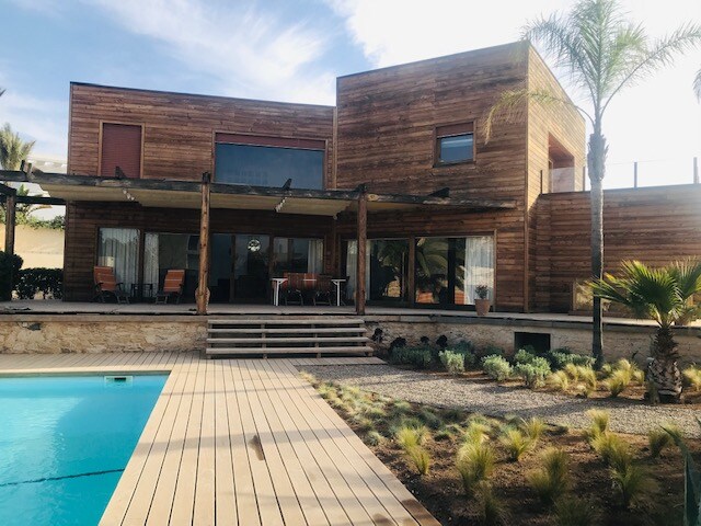 A wooden villa features large glass doors opening to a deck and a swimming pool. The outdoor space is landscaped with low shrubs and palm trees. A covered seating area is visible, providing shade, and the structure has multiple levels and windows, creating a bright and open environment.