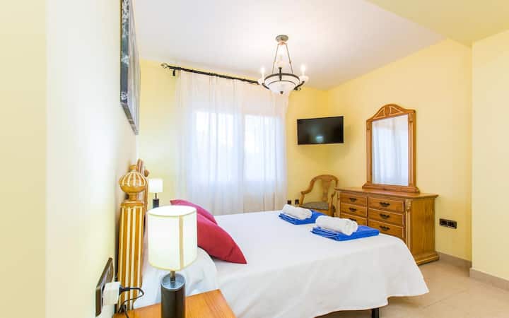 La cama es el elemento central de la habitación. Tiene una cabecera de madera tallada y está vestida con sábanas blancas y una colcha. Dos almohadas grandes y dos almohadas decorativas en tonos rojos añaden un toque de color y confort.