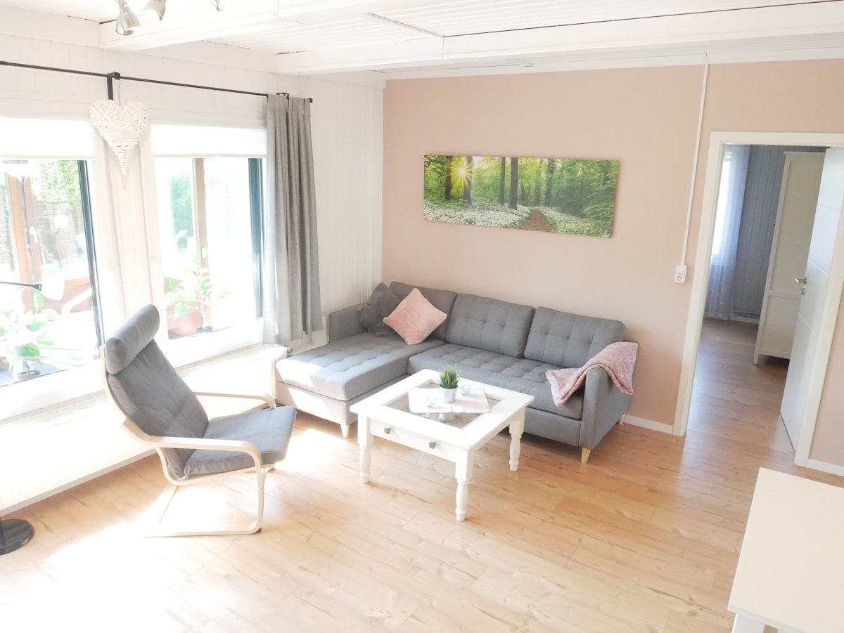 A light-filled living area features a gray sectional sofa paired with a cushioned armchair. A white coffee table occupies the center, with a small potted plant on top. Large windows allow natural light to illuminate the space, enhancing the warmth of the wooden flooring.