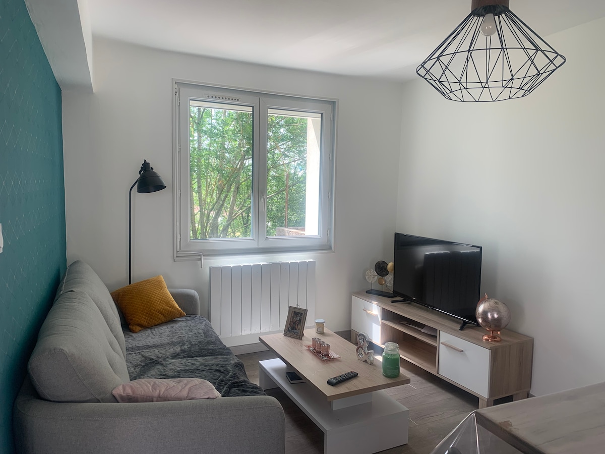 A light-filled living area features a gray sofa complemented by a yellow throw pillow. A modern lamp stands next to the sofa, while a flat-screen television is placed on a wooden media console. Natural greenery is visible through the large windows.