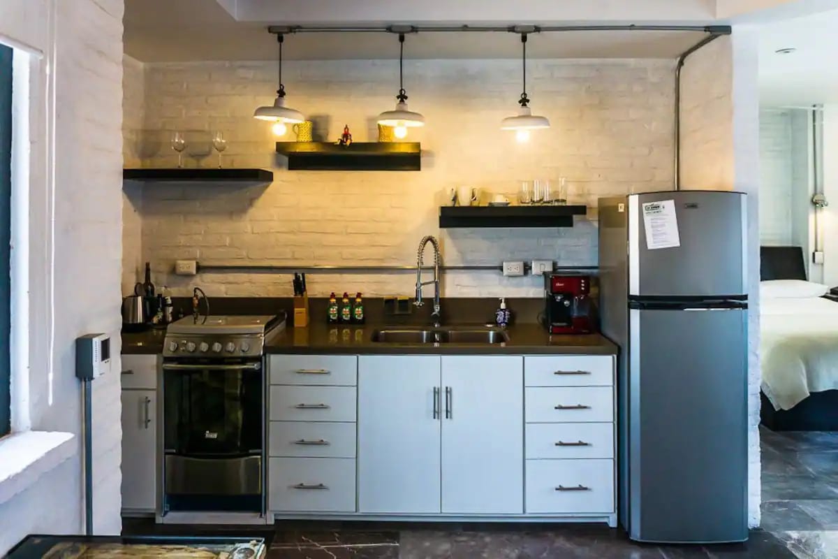 A functional kitchen area features modern appliances including a stainless steel refrigerator and a gas stove. Shelves hold glassware and decorative items. Task lighting hangs above the countertop, enhancing the warm ambiance. The design is complemented by a neutral color palette and sleek cabinetry.