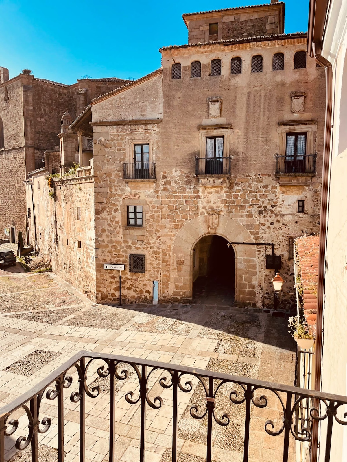 The image captures a historic stone building featuring multiple levels and a prominent archway. Decorative iron balconies accentuate the exterior, while sunlight bathes the structure, highlighting its textured walls. The surrounding cobblestone area adds to the sense of the charming neighborhood.