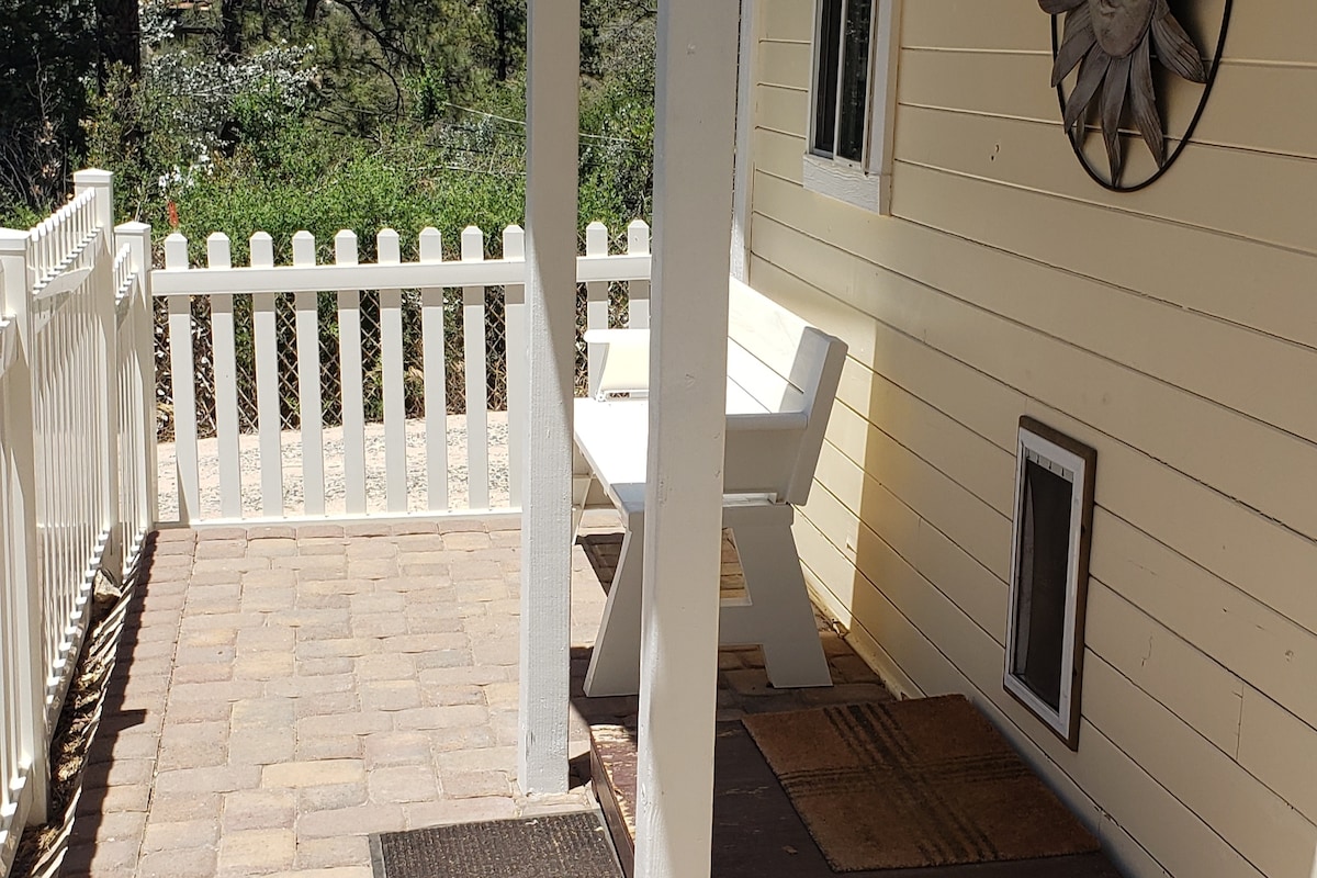 A covered porch area is highlighted by a white bench and a sun-themed wall decoration. Paved stones create a neat walkable surface, and a wooden fence with vertical slats provides a boundary, allowing natural light and views of the surrounding greenery.