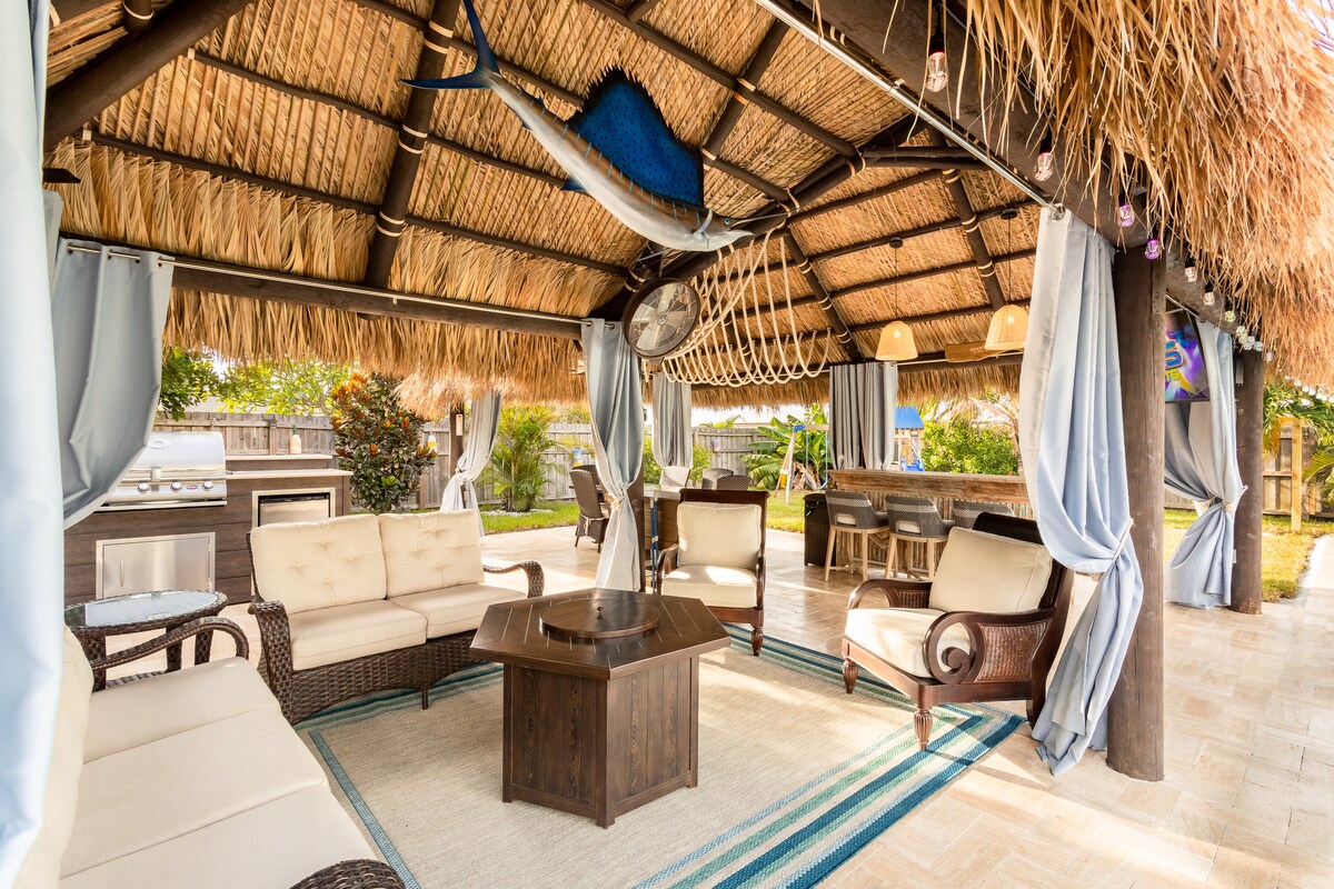 A spacious tiki hut features thatched roofing and comfortable seating arrangements beneath. High-quality outdoor furniture is paired with a solid wood bar, creating an inviting atmosphere. Decorative elements include curtains and light fixtures, while the lush backyard is visible in the background.