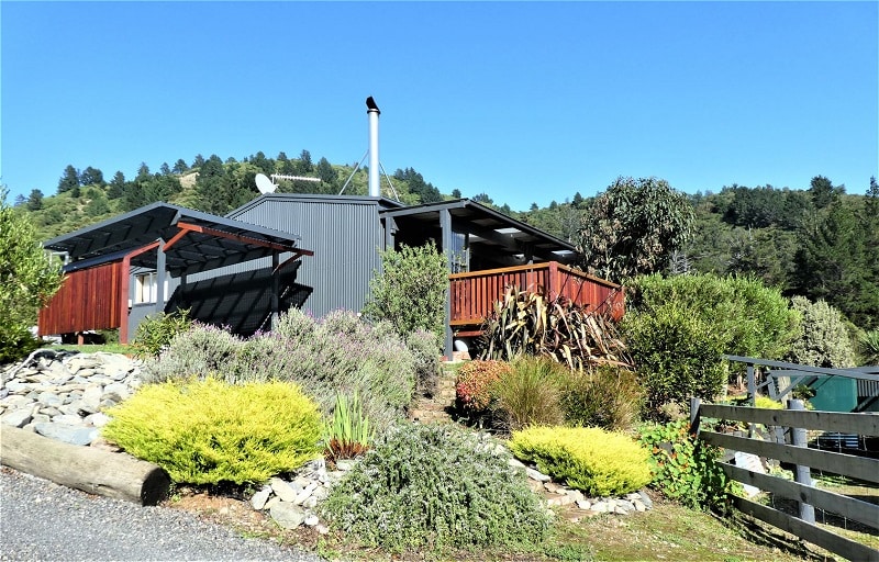 The exterior of a modern home is framed by lush greenery and neatly arranged stone pathways. A spacious deck with wooden railings is visible, alongside a stack of firewood. The surrounding landscape showcases a mix of shrubs and small trees against a clear blue sky.
