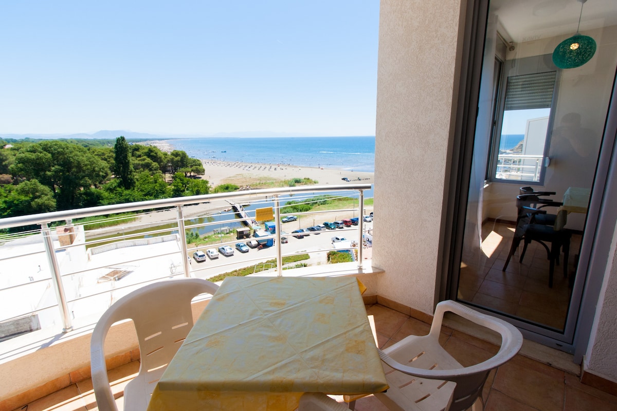 A balcony area features a small table and two white chairs, overlooking a scenic view of the sea and beach. The clear sky and gentle waves are visible in the background, with nearby green trees adding to the serene atmosphere.