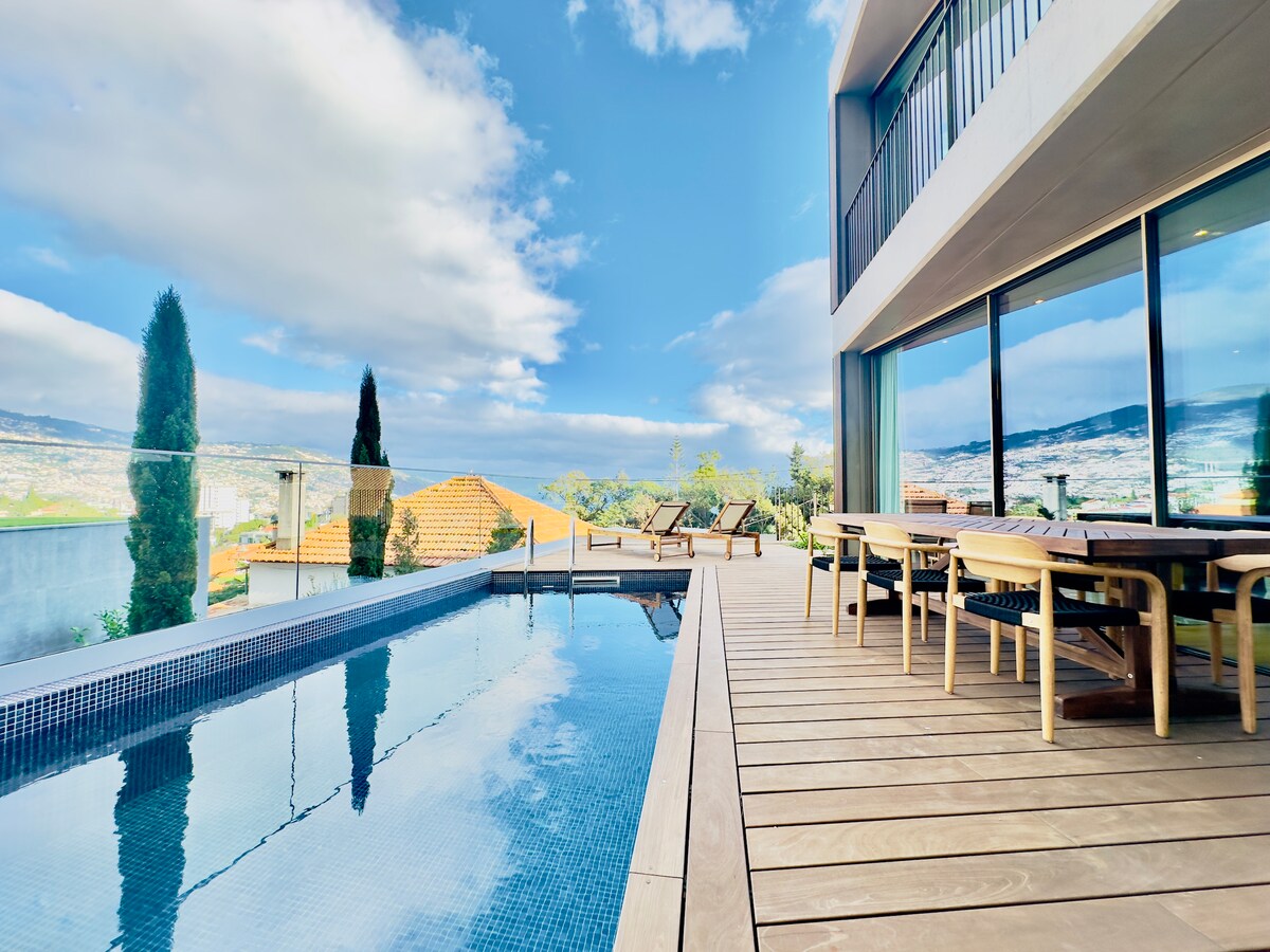 An outdoor terrace features a serene swimming pool reflecting the sky, surrounded by sun loungers and a dining area. The space is framed by tall greenery, with expansive glass windows offering views of the surrounding landscape.