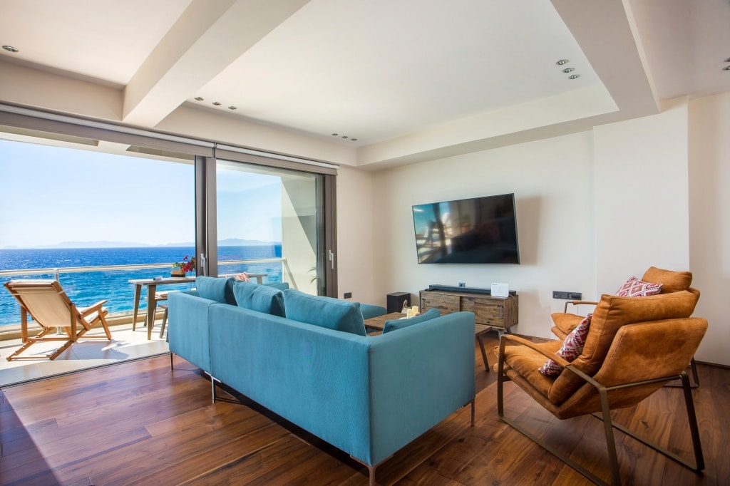 A modern living room features a large, comfortable sofa and two accent chairs positioned toward a wall-mounted television. Floor-to-ceiling glass doors open to a balcony, revealing expansive sea views. Natural light enhances the space, highlighting the warm wood flooring.
