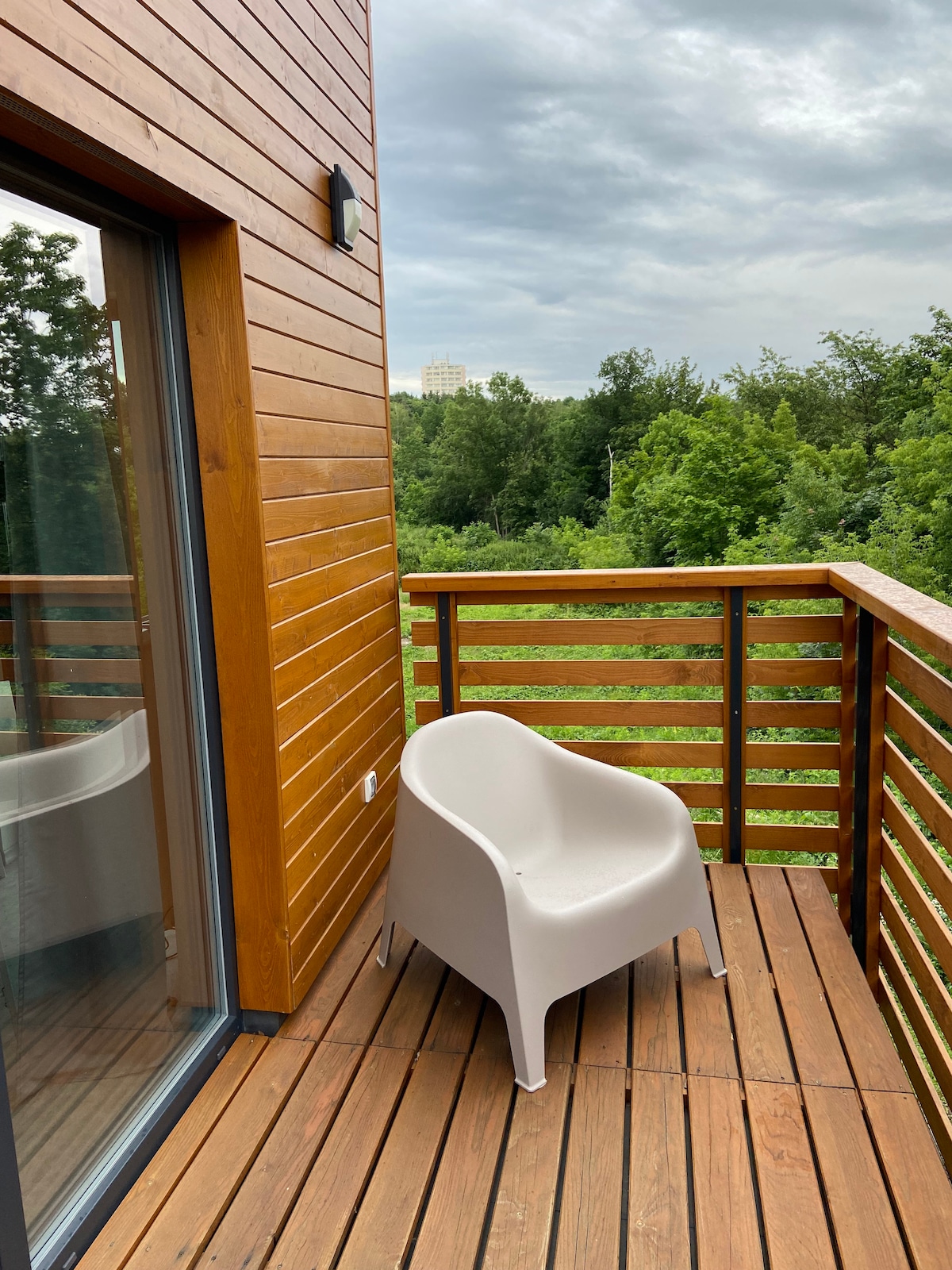 A private terrace is depicted, featuring a sleek, white plastic chair positioned on a wooden deck. The railing provides a view of lush greenery beyond, with a partially cloudy sky overhead. Natural wood accents are visible on the walls, contributing to a modern aesthetic.