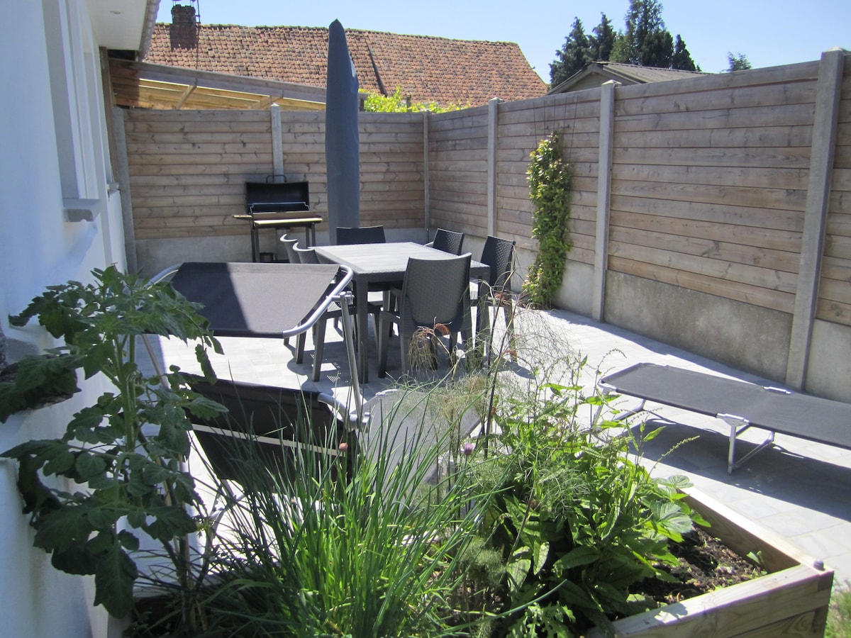 An outdoor terrace space is shown, featuring a dining table surrounded by several chairs. Sun loungers are positioned nearby. A barbecue grill is visible in the corner. The area is enclosed by wooden fencing and includes a small garden with herbs and plants.