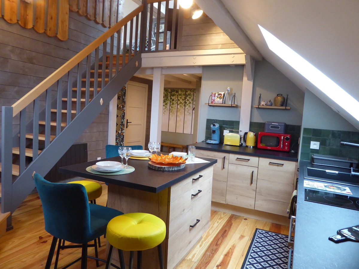 A bright and open kitchen area features a large island with seating for four, complemented by modern appliances in muted tones. Wooden beams and a staircase add character to the space, while large windows provide natural light.