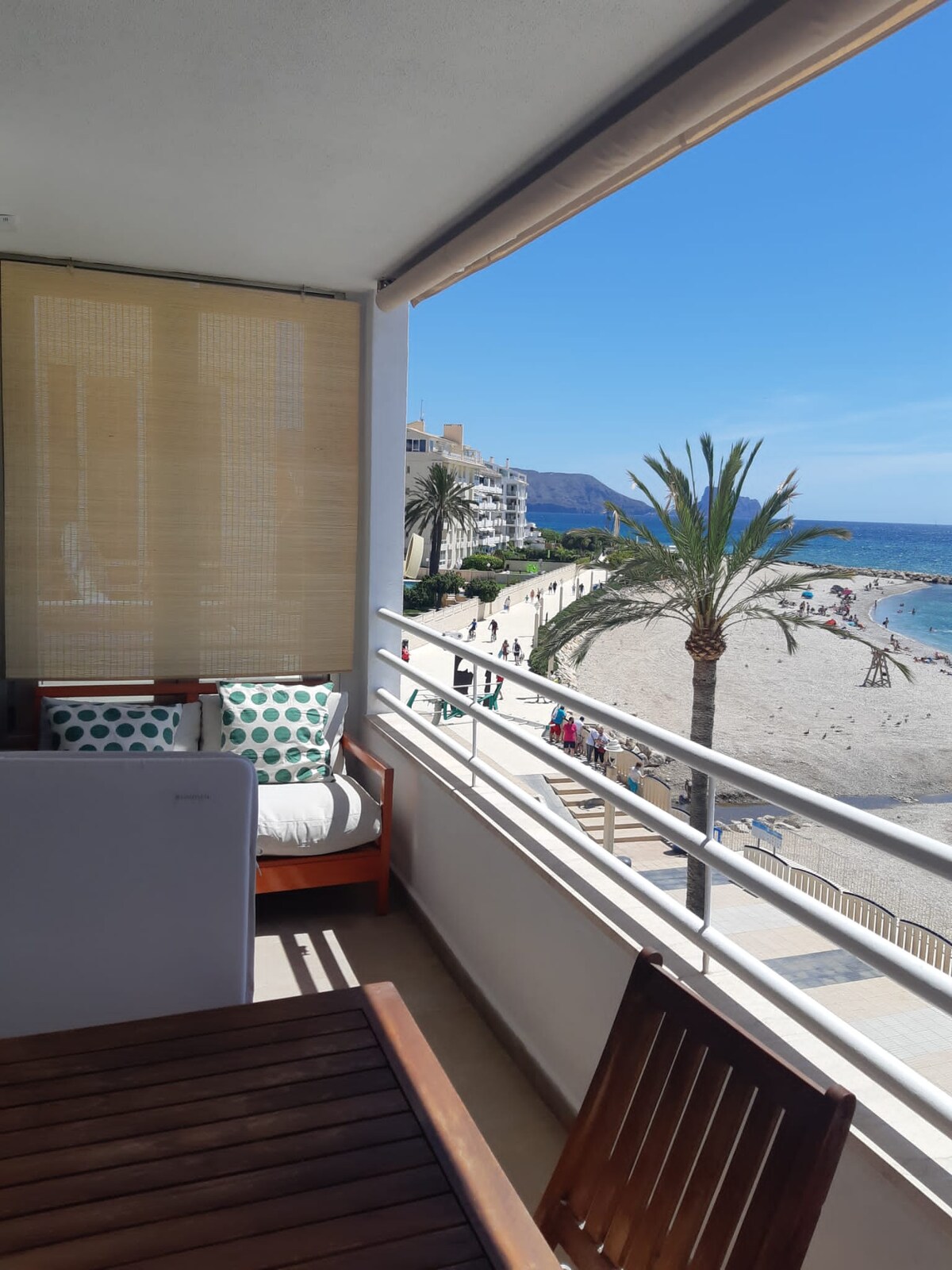 A private balcony offers views of the nearby beach and palm trees. Outdoor seating is provided, featuring cushioned chairs and a wooden table. The surrounding area includes sandy shores and a promenade, with people enjoying the beachfront activities under a clear blue sky.
