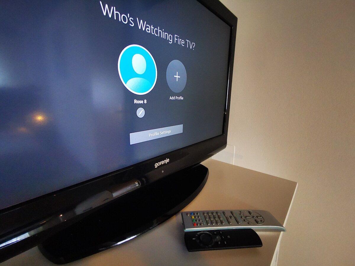 A flat-screen TV is displayed on a light-colored console, showing a Fire TV interface prompting for user profiles. A remote control sits next to the television, allowing for easy access to entertainment options.