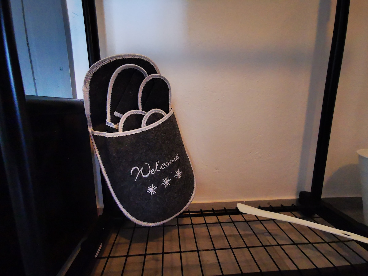 A pair of cozy grey slippers, featuring the word 'Welcome' in white embroidery, is displayed on a black wire shelf. The shelf is situated in a well-organized corner, contributing to a homely ambience.