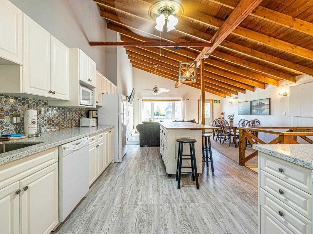 The kitchen area features white cabinetry and a mosaic backsplash, complemented by a spacious countertop. An open layout connects the kitchen to a dining area, illuminated by natural light from large windows. Exposed wooden beams create visual interest on the ceiling.