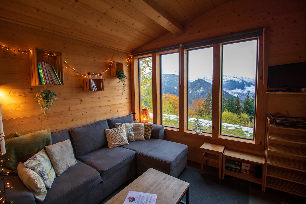A cozy living area is presented, featuring a spacious gray sectional sofa adorned with decorative cushions. Large windows offer a panoramic view of the surrounding mountains, while warm lighting and wooden accents create a welcoming atmosphere.