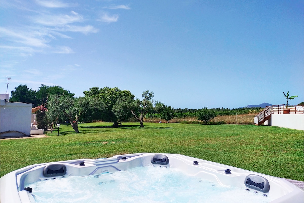 A jacuzzi sits in the foreground, surrounded by a well-kept lawn. In the background, various trees, including olive trees, create a serene landscape, while the sky remains clear and blue. A distant view of rolling hills can be seen beyond the greenery.