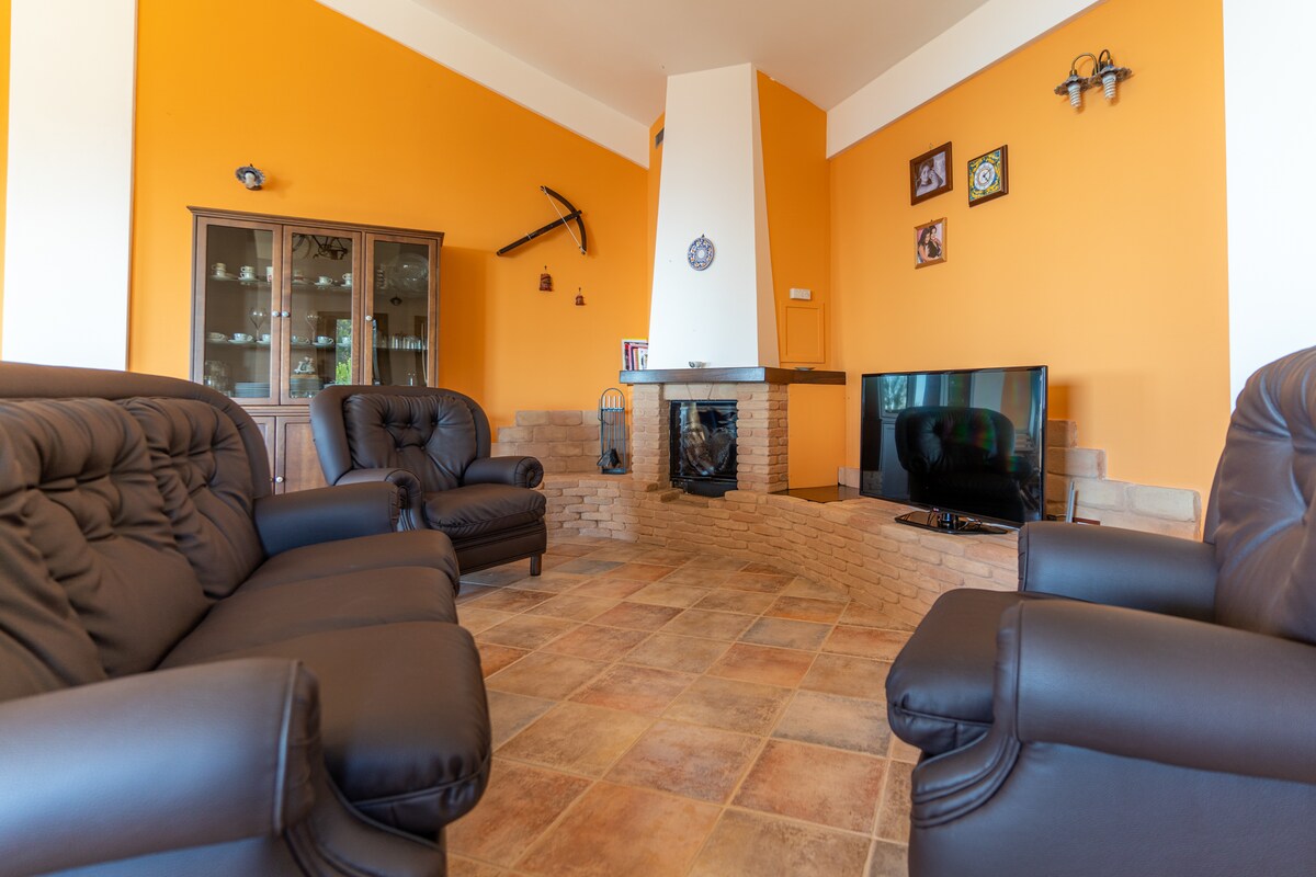 A living area features comfortable black leather seating arranged around a low stone fireplace. The room is enhanced by warm orange walls and a television placed on a small cabinet. Photographs are displayed on the walls, adding character to the space.