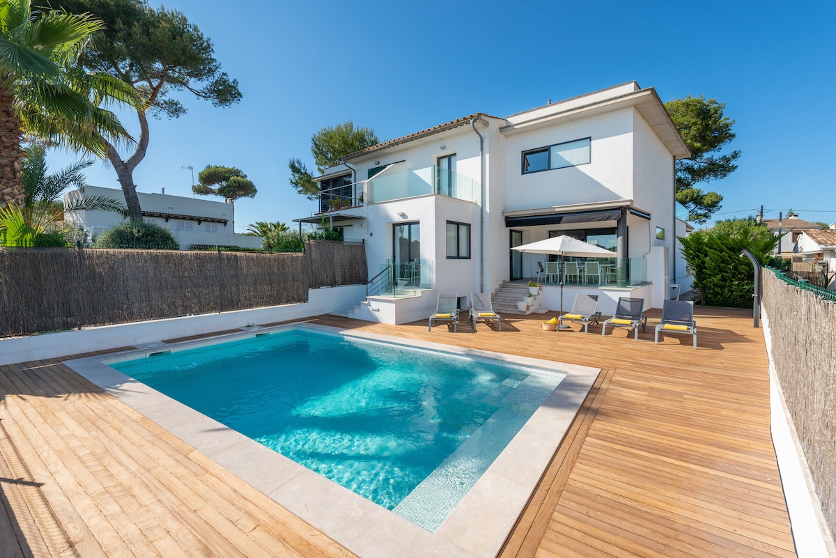 The exterior of the villa is showcased, featuring a spacious swimming pool surrounded by wooden decking. Comfortable sun loungers are positioned nearby, along with a shaded sitting area. The modern two-story structure includes large windows, allowing plenty of natural light to enter.