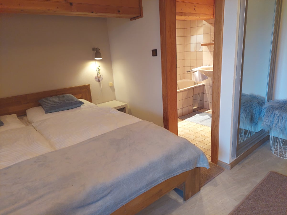 A simple bedroom features a spacious bed with a light blue blanket. Soft lighting is provided by a wall-mounted lamp. An open doorway leads to a bathroom with tiled walls and a bathtub, while fluffy stools are positioned near a mirror.