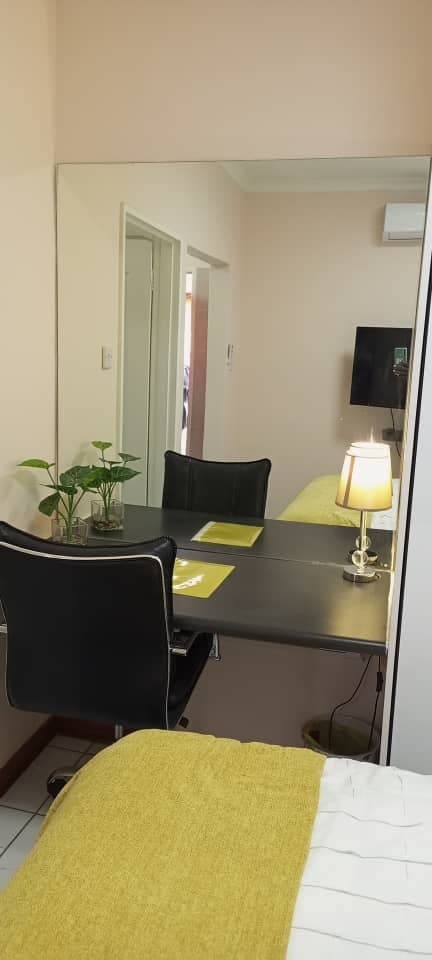 A sleek desk with a black chair is positioned against a light wall, illuminated by a small lamp. A potted plant adds a touch of greenery. A mirror reflects the workspace, enhancing the room's spacious appearance. Soft bedding in the foreground is draped in a yellow cover.