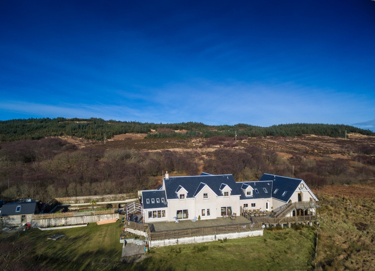 An aerial view of a spacious house surrounded by a lawn, with multiple sections and a variety of roof styles. The landscape includes rolling hills in the background under a clear blue sky, enhancing the property's peaceful setting.