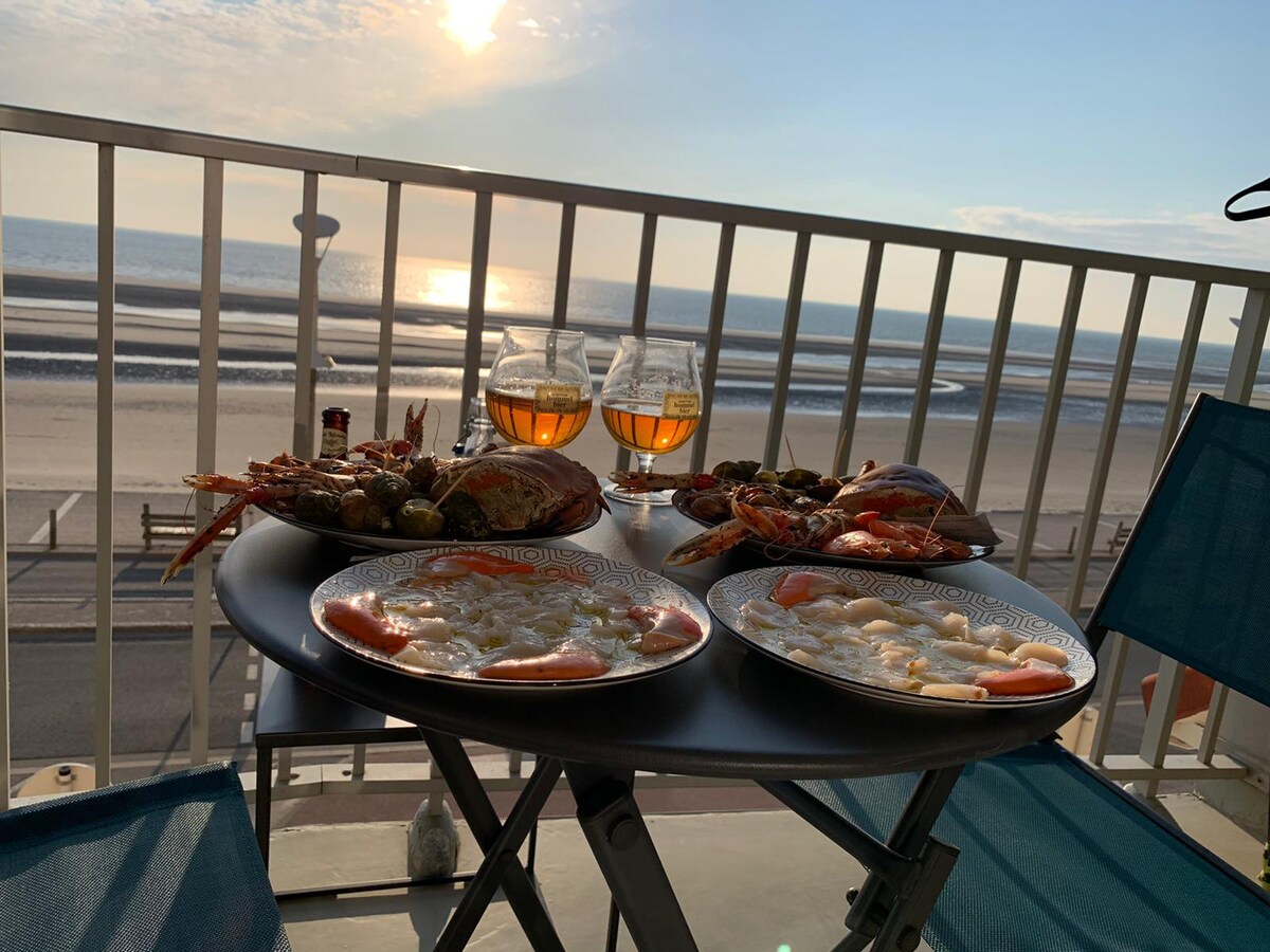 A balcony table is set for two, featuring plates of seafood and drinks, with glasses of amber liquid. The ocean is visible in the background, reflecting the warm glow of the setting sun, while a calm beach stretches out below.