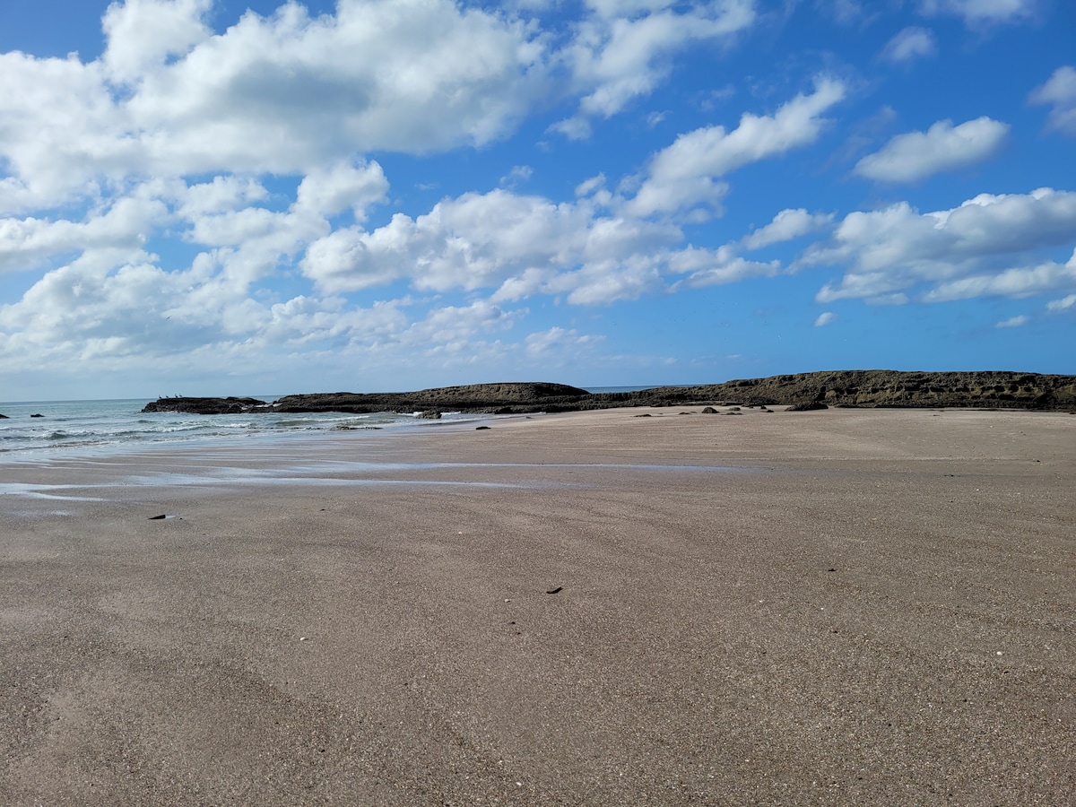 A sandy beach extends towards the horizon, under a sky filled with clouds. Gentle waves lap at the shore, while a rocky formation can be seen in the distance. The expansive shoreline offers a sense of tranquility and openness.