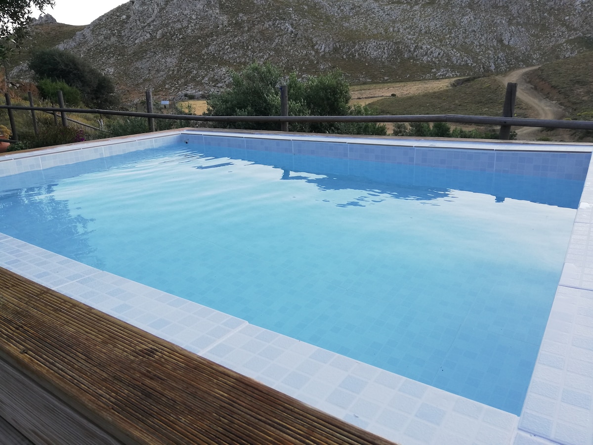 A clear swimming pool is set on a wooden deck, reflecting the surrounding landscape. The water appears calm and inviting, with gentle ripples visible on the surface. Dry grass and distant hills create a serene backdrop.