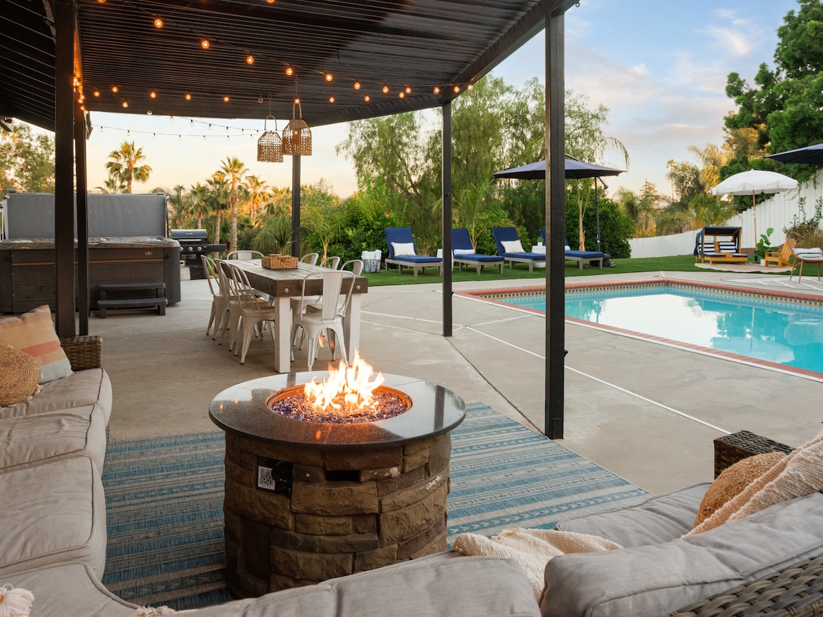 An outdoor seating area is equipped with a sectional couch surrounding a stone fire pit, with flames flickering invitingly. A large dining table offers ample seating. The heated pool glistens in the background, surrounded by lounge chairs and shaded umbrellas.