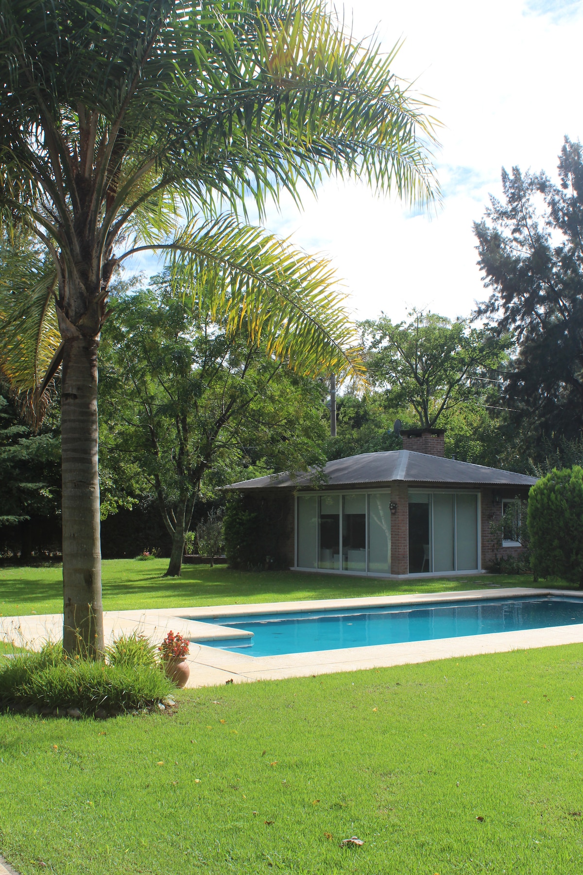 An outdoor area features a private swimming pool surrounded by lush greenery. A well-maintained lawn is complemented by a cozy poolside terrace. In the background, a serene guesthouse is partially hidden by trees, creating a tranquil and inviting environment.