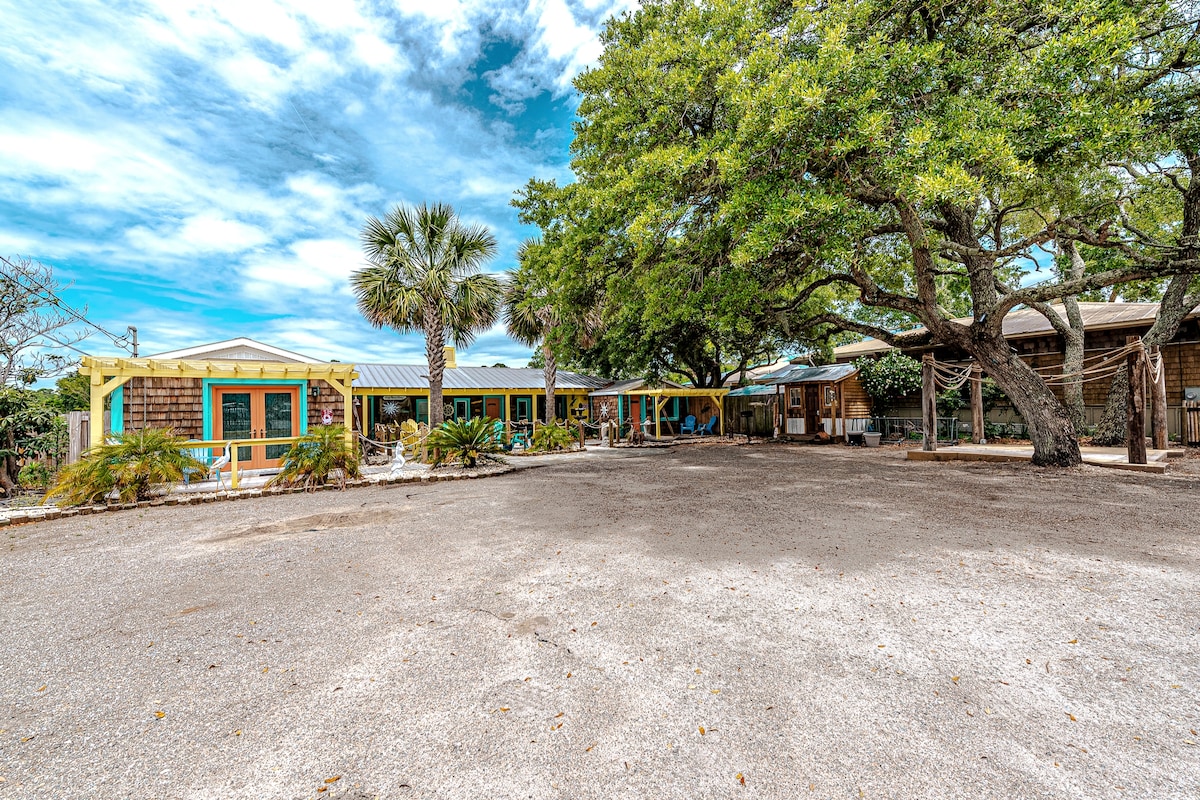 A spacious outdoor area is surrounded by vibrant trees and features multiple bungalow-style units with colorful trim. A sandy path leads up to the front porches, while additional seating areas are visible under the shade of large trees, providing a relaxed, nature-filled environment.