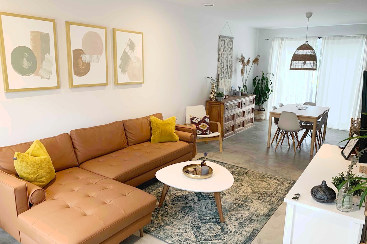 A modern living space is presented, featuring a large, tan leather sectional sofa adorned with a yellow pillow. An abstract art piece is displayed above the couch. A round coffee table sits at the center, with a woven bowl. Light pours in through sheer curtains, illuminating the area.