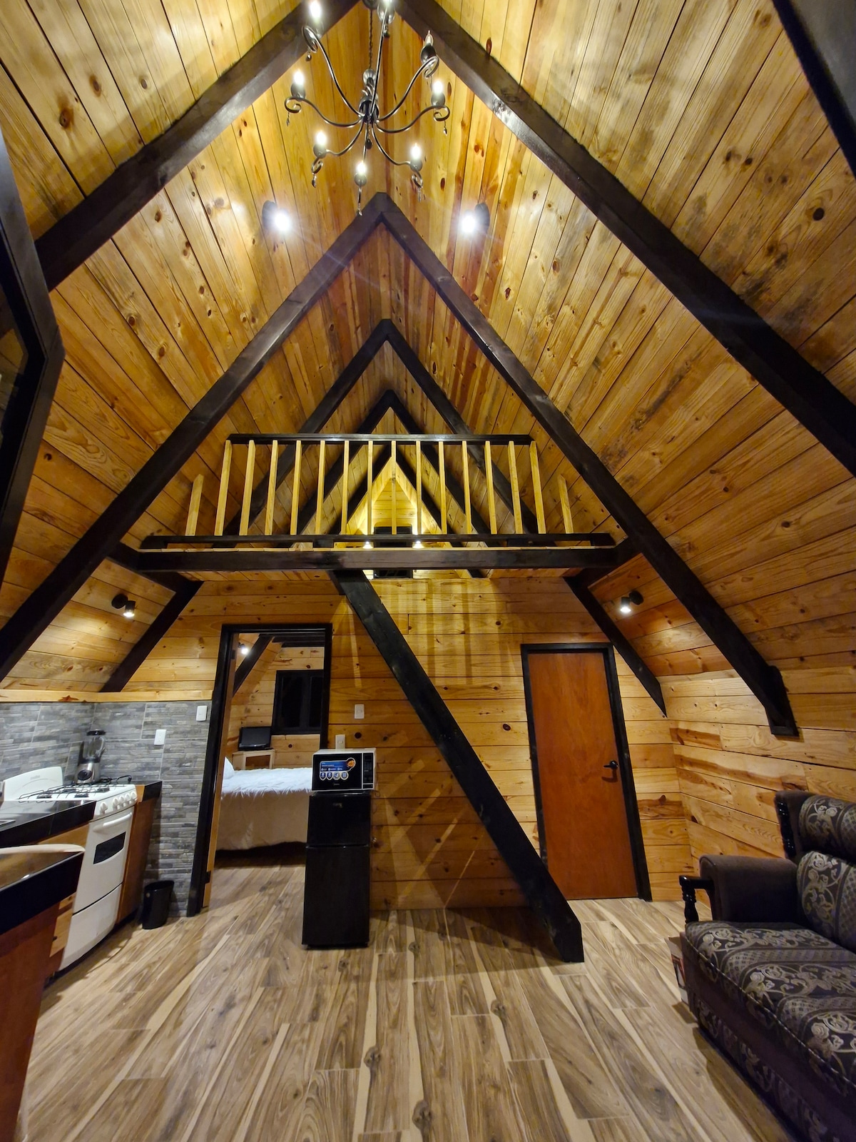 An inviting cabin interior features a wooden ceiling with exposed beams and a loft area. The walls are lined with warm wood paneling. A cozy seating area is visible, adjacent to a small kitchen area equipped with essential appliances.